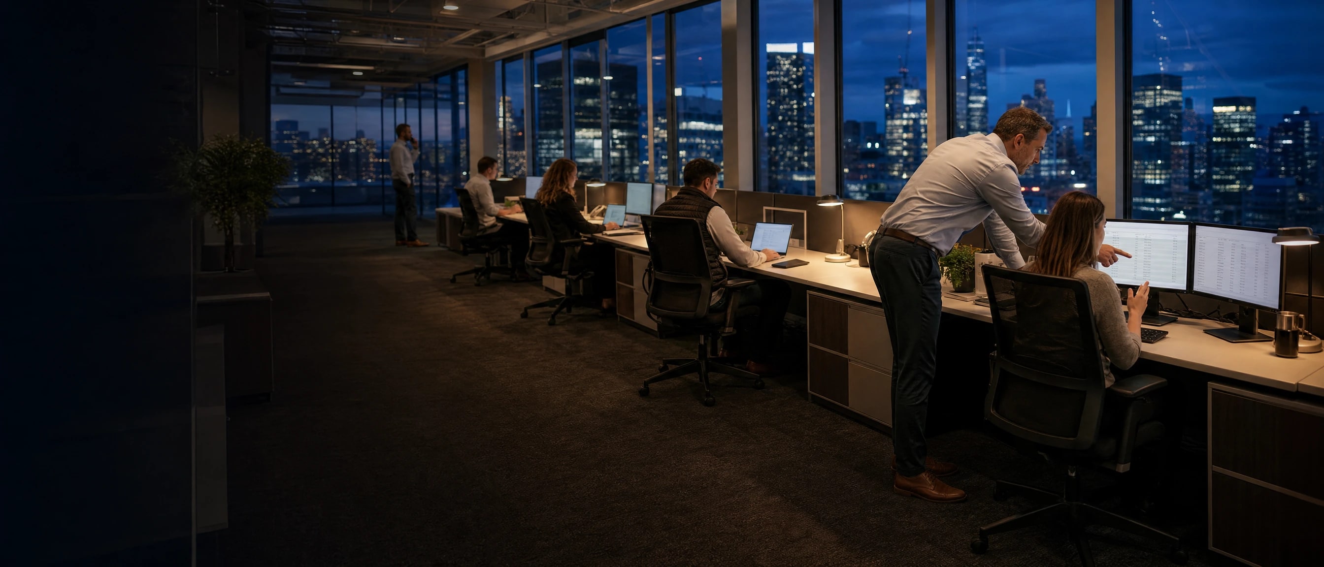 A producer and account manager working the renewal list together on the agency floor, the team along the windows at blue hour